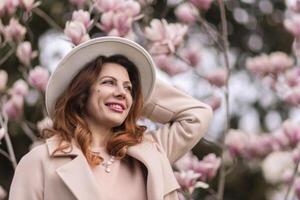 Woman magnolia flowers, surrounded by blossoming trees., hair down, white hat, wearing a light coat. Captured during spring, showcasing natural beauty and seasonal change. photo