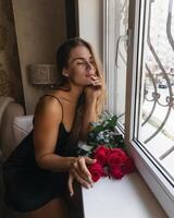 A woman in a black dress is sitting in front of a window with a bouquet of red roses. photo