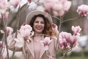 Woman magnolia flowers, surrounded by blossoming trees., hair down, white hat, wearing a light coat. Captured during spring, showcasing natural beauty and seasonal change. photo