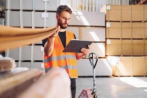 Holding stick and looking into tablet. Storage worker is in the warehouse with bunch of products photo