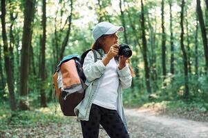 Standing and holding camera in hands. Girl is in the forest at summer day time discovering new places photo