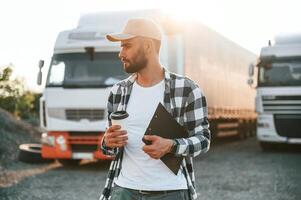 With cup, of drink and notepad. Young truck driver is with his vehicle at daytime photo
