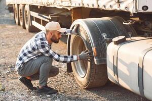 Working with damaged tire. Young truck driver is with his vehicle at daytime. photo