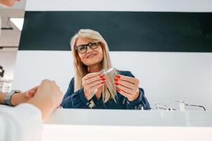 Sitting by the table.. Woman choosing glasses in the store and getting help by assistant photo