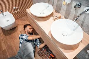 Laying ,down on the ground, fixing the sink. Plumber in uniform is indoors at home photo