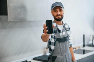 Holding, smartphone, in hand. Plumber in uniform is indoors at home photo