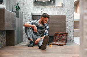 On the floor, with case of equipment.. Plumber in uniform is indoors at home photo