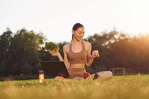 Using l,aptop. Holding vegetables. Young woman in yoga clothes is outdoors on the field photo