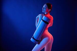 Standing ,with yoga mat. Young woman in sportive clothes is in the studio with neon lights photo