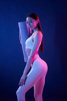 Ready for exercises.. With yoga mat. Young woman in sportive clothes is in the studio with neon lights photo