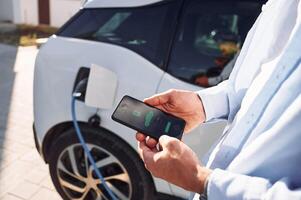 Process ,of charging .is showing on smartphone. Close up view of man with his electric car photo