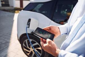 Process, of charging is showing on smartphone. Close up view of man with his electric car photo