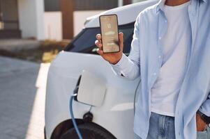 Process ,of charging is showing on smartphone. Close up view of man with his electric car photo