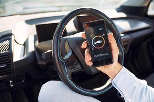 Process of charging is showing on smartphone. Close up view of man with his electric car photo