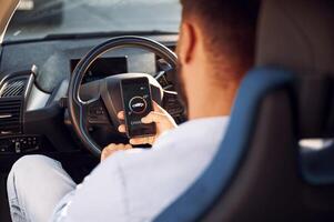 Sitting inside the vehicle, monitoring charging process by using smartphone. Young stylish man is with electric car photo
