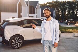 Standing a,nd holding the keys. Young ,stylish man is with electric car at daytime photo