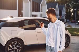 Standing a,nd holding the ,keys. Young stylish man is with electric car at daytime photo