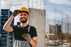 In hard hat, with, notepad. Man in uniform is working on the construction site photo