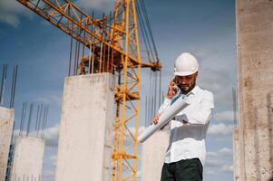 Talking by phone and holding document in hands. Man in uniform is working on the construction site photo