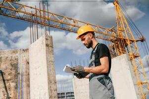 Standing against yellow crane, and holding notepad with documents. Man in uniform is working on the construction site. photo