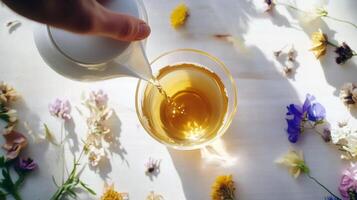 Pouring herbal tea into glass cup surrounded by colorful flowers on white table photo