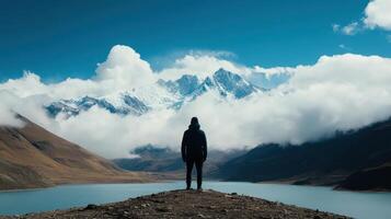 Adventurous Hiker Standing on Mountain Peak Overlooking Serene Lake and Snow-Capped Mountains Under a Clear Blue Sky photo