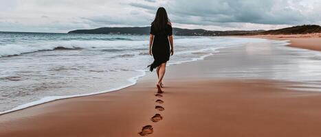 A woman walks along a sandy beach, leaving footprints in the wet sand as waves gently lap at the shore under a cloudy sky. photo