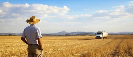 Economic Inequality Concept. A farmer in a straw hat watches a truck on a vast golden field under a blue sky with scattered clouds. photo
