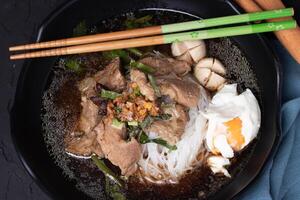 flavorful bowl of beef noodle soup with tender beef slices, soft boiled egg, and fresh herbs, garnished with fried garlic and served with chopsticks photo