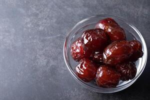 glass bowl filled with glossy, red candied dates sits on dark, textured surface, showcasing their rich color and shiny appearance photo