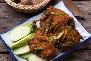 Spicy fried fish garnished with herbs and served with fresh cucumber slices on rustic wooden table. dish is presented on rectangular plate, highlighting its vibrant colors and textures photo