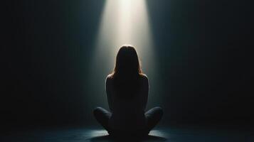 woman sits alone in dark room, illuminated by single beam of light, creating contemplative and serene atmosphere photo
