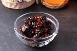 glass bowl filled with dark, glossy preserved fruit sits on dark surface, showcasing rich and sticky texture. background features blurred basket with flowers, adding rustic touch to scene photo