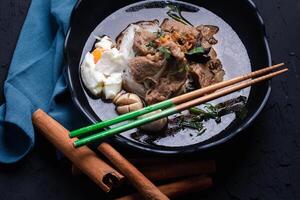 bowl of flavorful beef noodle soup with poached egg and herbs is served in black dish, accompanied by chopsticks and blue napkin on dark textured surface photo