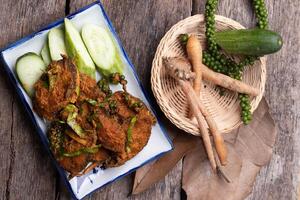 plate of spicy fried fish cakes garnished with fresh cucumber slices and green peppercorns, accompanied by basket containing fresh fingerroot and cucumber, all set on rustic wooden table photo