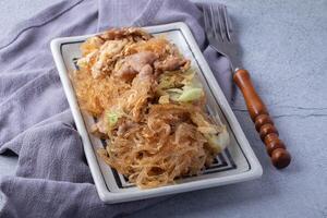 plate of stir fried glass noodles with pork and cabbage is served on rectangular dish, placed on gray cloth with wooden handled fork beside it photo