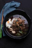 bowl of flavorful beef noodle soup with tender beef slices, poached egg, and fresh vegetables, garnished with herbs, served in black bowl on dark textured surface photo