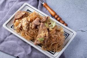 rectangular plate of stir fried glass noodles with pork, cabbage, and egg on gray cloth. dish is served on textured gray surface, accompanied by wooden handled fork photo
