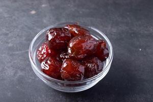 glass bowl filled with glossy, dark red candied dates sits on dark textured surface, showcasing their rich color and sticky texture photo