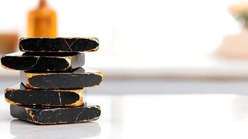 Stacked black soap bars with golden edges on a bright countertop, with blurred background elements photo
