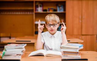 Emotional cute boy in glasses and a white shirt at a school desk with textbooks photo