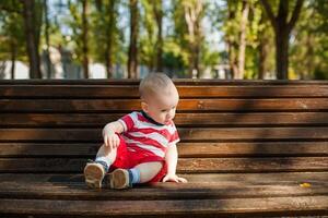 Baby boy in red shorts and striped shirt sitting on a wooden bench in summer warm park photo