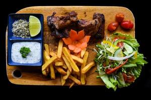 Grilled smoked pork ribs with french fries and sauces on wooden board on black background side view photo