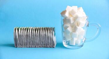 Many tea bags in a row next to a transparent glass mug full of white refined sugar on blue background photo