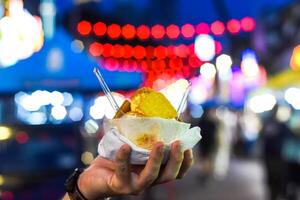 Hand holding coconut and mango ice cream in coconut shell against lights of Jalan Alor street food market in Kuala Lumpur photo