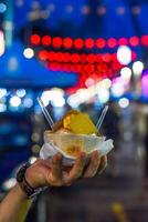 Hand holds coconut and mango ice cream in coconut shell against lights of Jalan Alor street food market in Kuala Lumpur photo