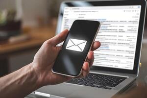 Hand holds smartphone displaying an envelope icon, with a laptop in the blurred background photo