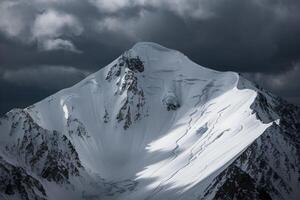 Snow covered mountain peak with dark clouds and sunlight highlighting rugged terrain photo