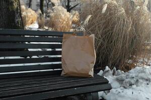 A paper craft bag with Takeaway food products groceries stands on a wooden bench in a winter sunny day. Fast delivery photo