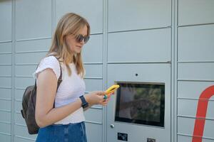 Cheerful young travel 30s woman using smart phone while standing near automatic lockers. New technologies in delivery service, self picking photo